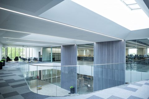 Photo of an open office space, in the foreground in a balcony overlooking an entrance area, in the background and seperated with a glass partition is an office area. The flooring is tiled carpet.