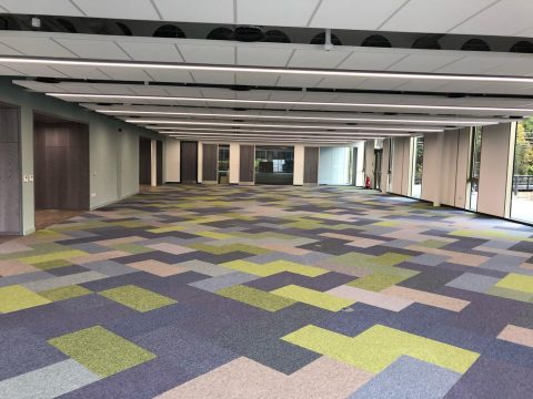 An image of a large room with several doors and windows. The flooring consists of tiled carpet in a range of different colours