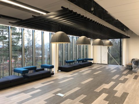 Photo of a seating area in front of large glass windows looking out to trees and a road. IN front of the windows are 2 black sofas, the flooring is a varied pattern of light and dark wood strips.